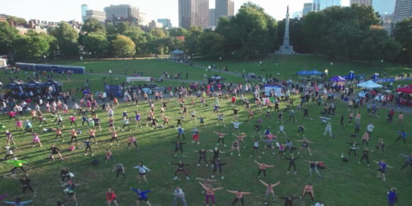 Aerial view of a large group of people exercising in a park. Many are spread out on the grass, performing stretches and workout moves. Tents and trees are visible in the background.